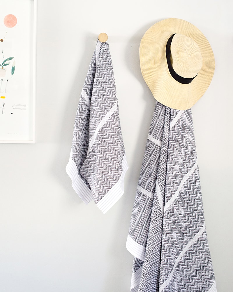 Mungo Tawulo Cotton Towels in Thunder Grey, hanging on wall with a hat. Textile woven from all natural fibres at our mill in Plettenberg Bay.