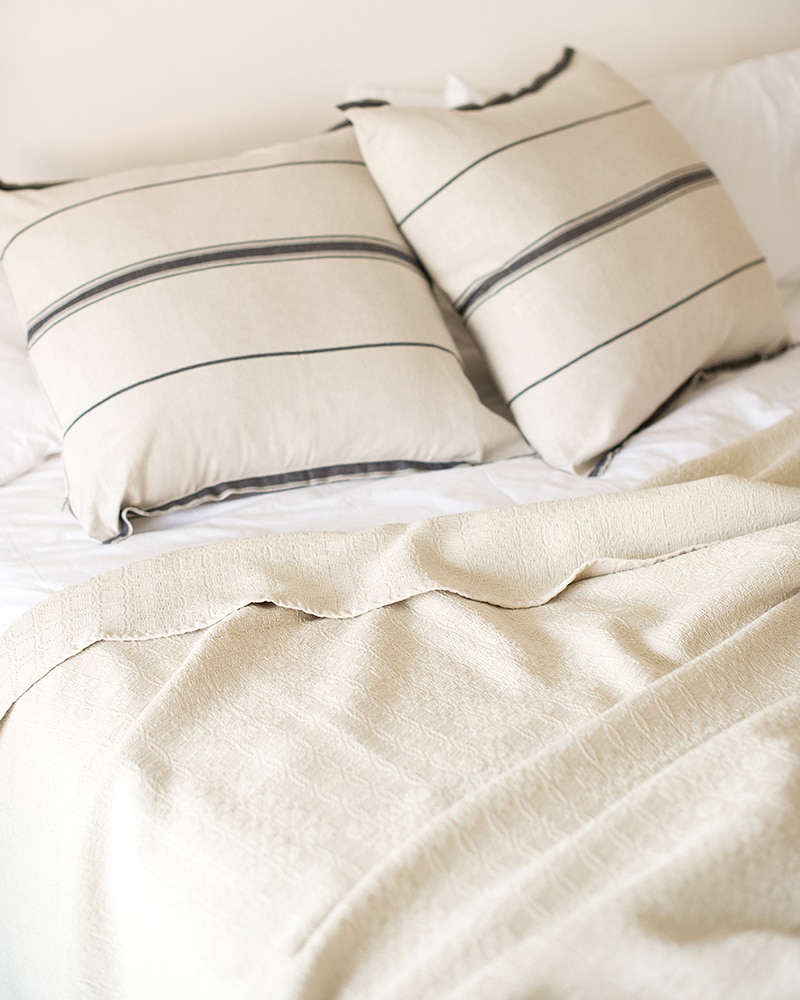 The Mungo Cotton Cottonfields Blanket Throw in Stone and the Linen Lisburn Cushion Cover in French Navy in a bedroom scene.