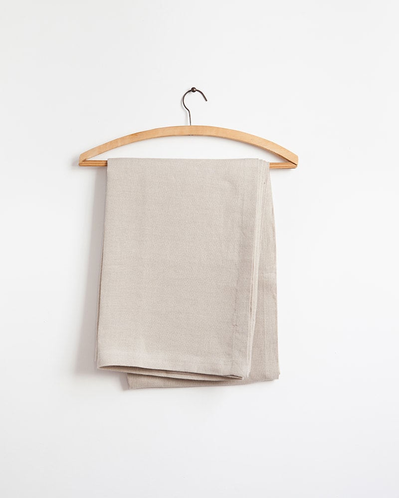 The Mungo Linen Table Cloth in Natural displayed on a wooden hanger. Made with natural fibres at the mill in Plettenberg Bay.