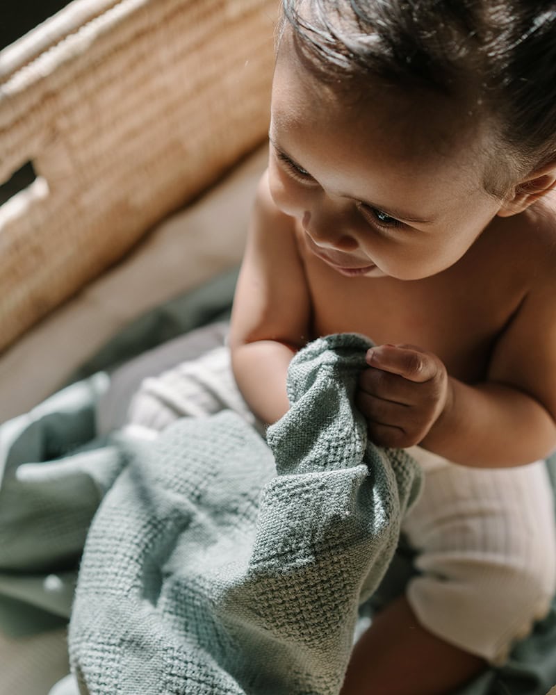 Baby sitting up holding a 100% Organic Cotton Mungo Cot Baby Blanket.