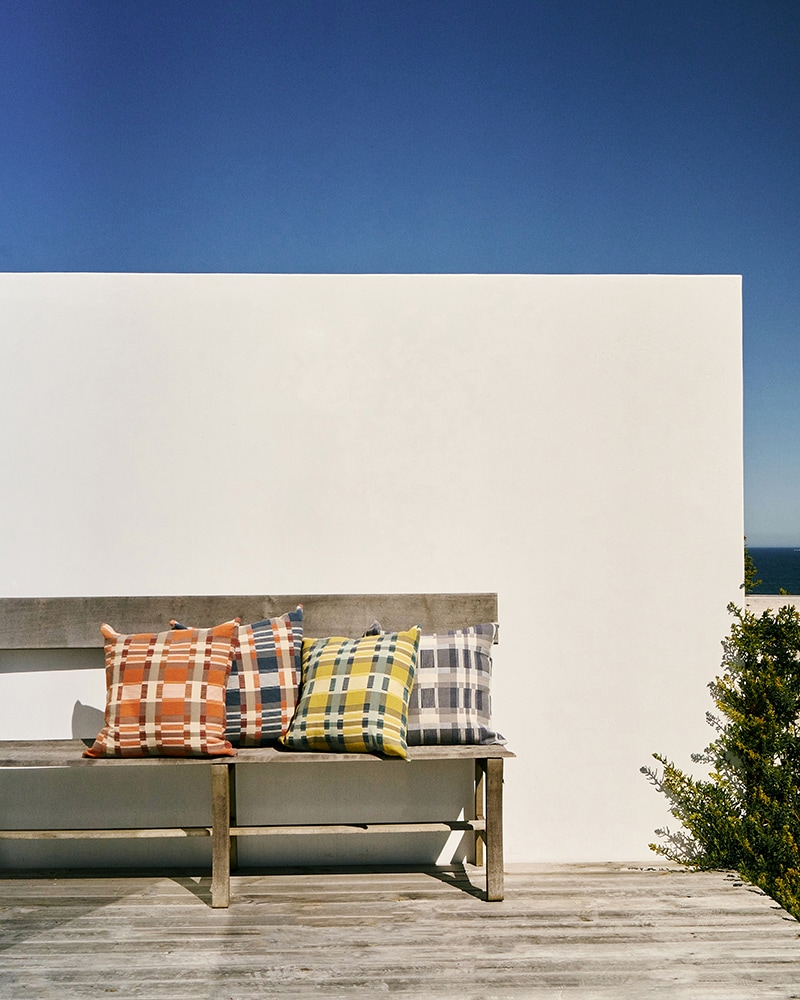 The Mungo Skipping Block Cushion Covers set on a bench in front of a walled and sky backdrop