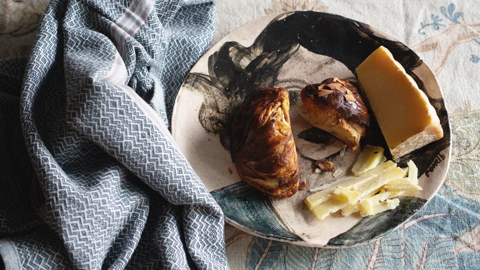 A table set with a tablecloth and a Mungo Boma Kitchen Cloth, with a plate on which sits a toasted cardamom bun and a wedge of delicious cheese.