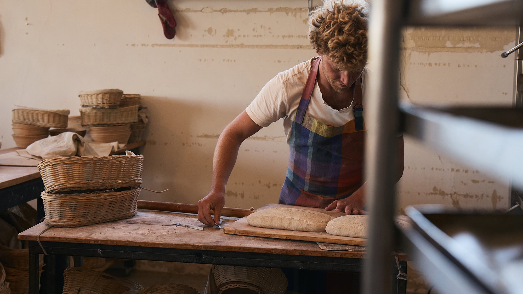 Banner shot of Brett Nortier behind baking trays, readying bread for his bakery and wearing the Mungo Alles apron in Iris.